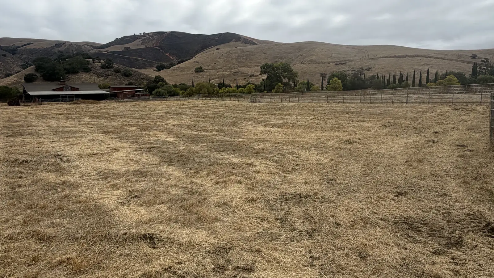 Pasture mowed short beside a pipe fence and ranch barns with dry hills behind, brush mowing