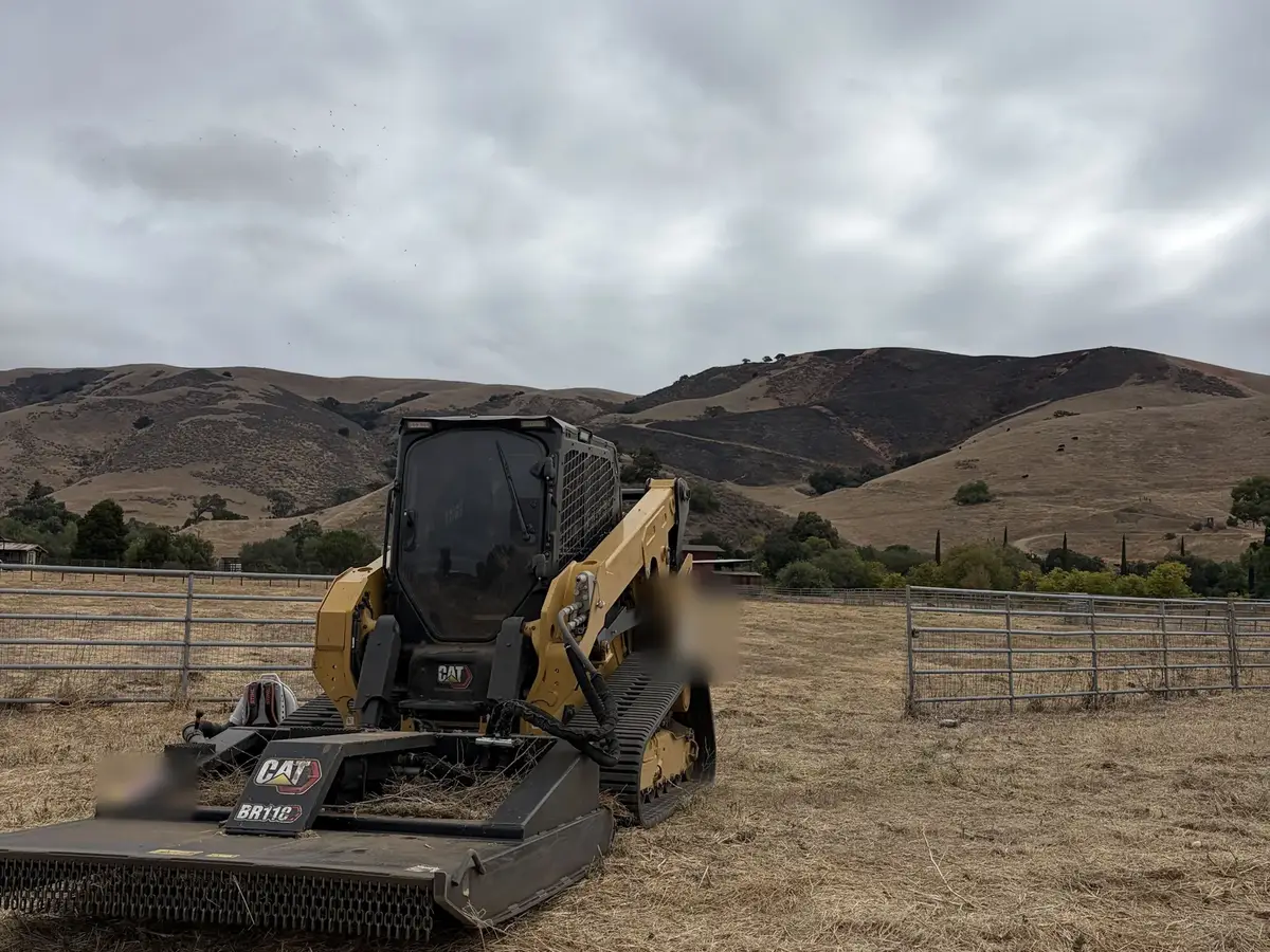 Compact track loader with a rotary brush mower parked in a mowed pasture with dry hills behind, weed abatement mowing